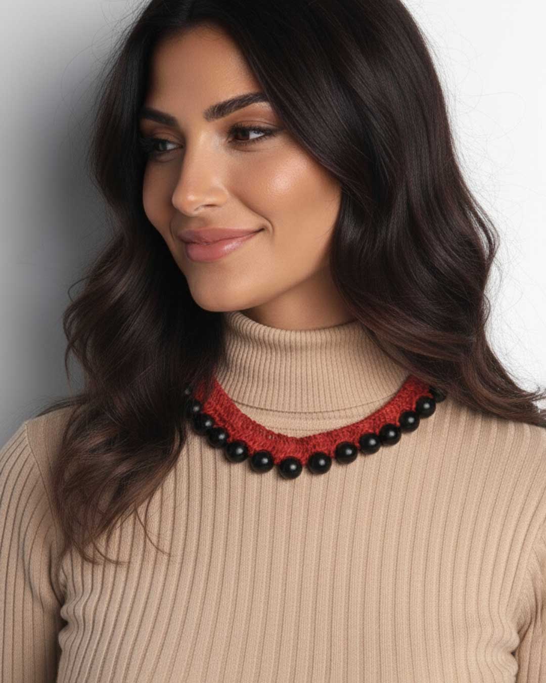 Woman wearing a beige sweater and black and red beaded necklace against a neutral background