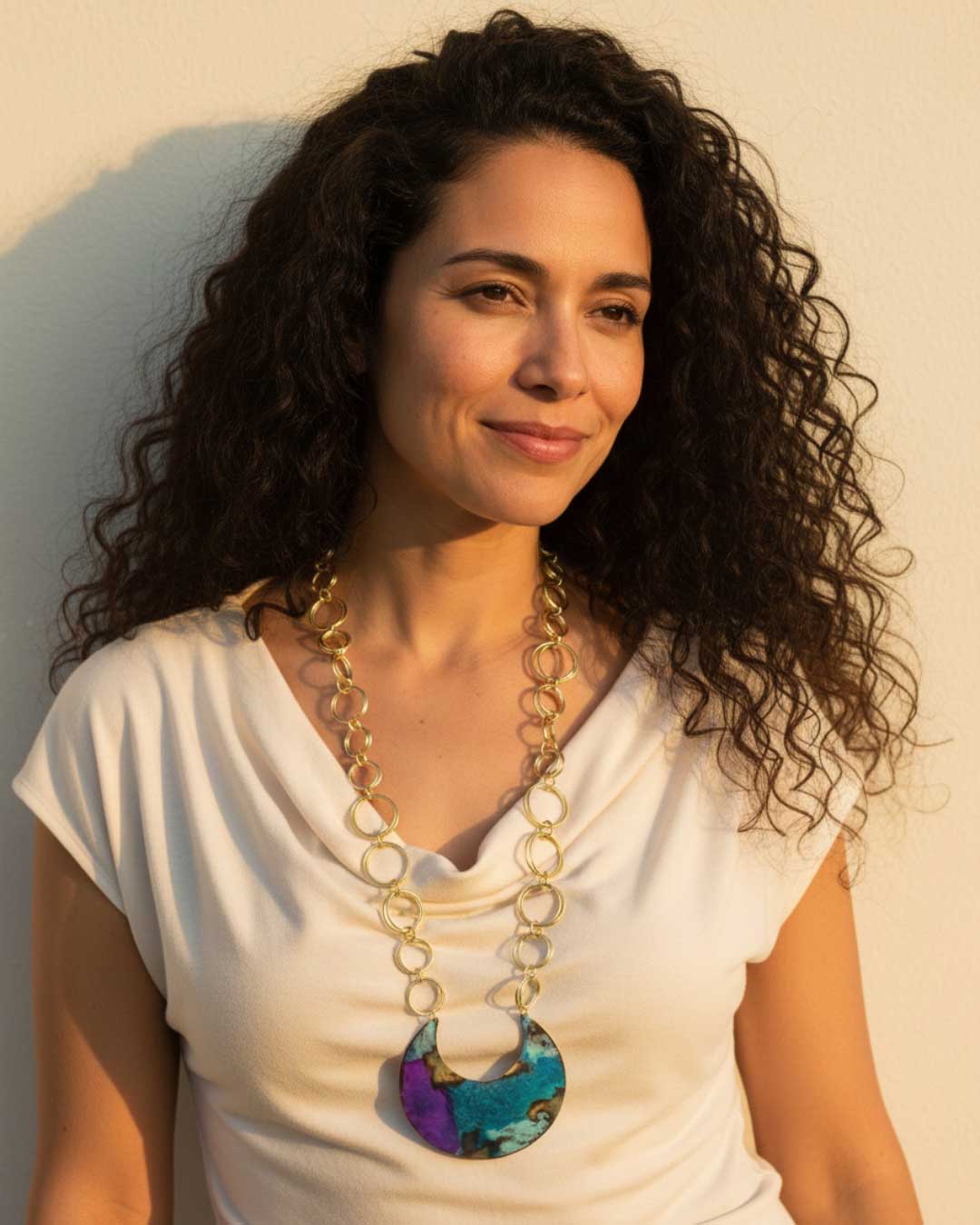 Hand-patinated brass crescent pendant necklace worn by a model in a white top photographed outdoors in warm golden light