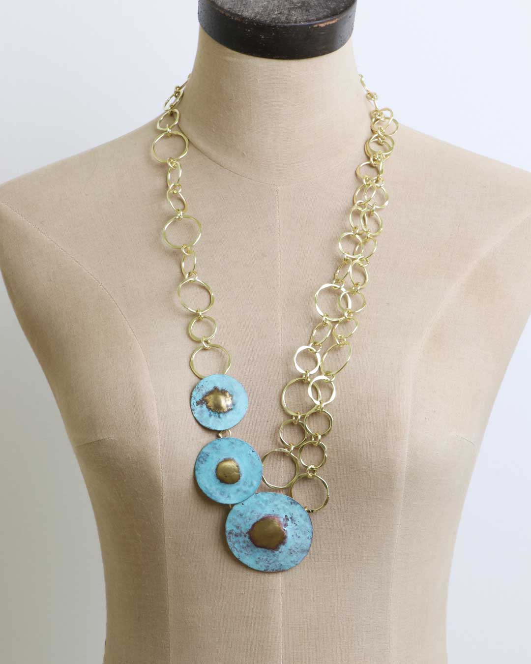 Hand-forged brass disc blue patina necklace on a beige dress form showing the drape and scale of the three-disc pendant on ring chain