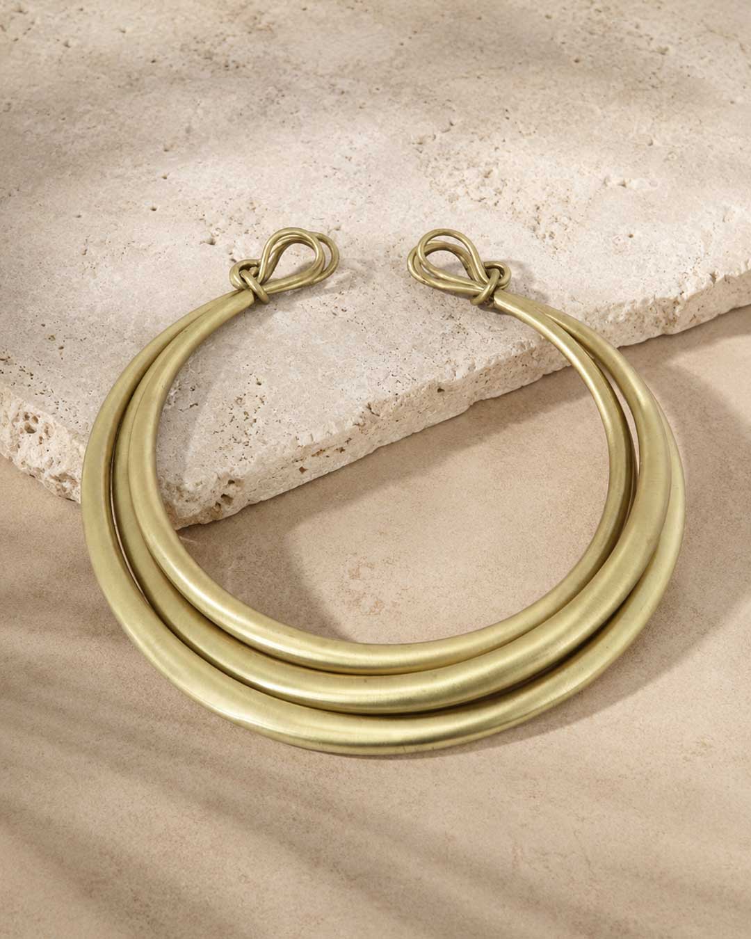 Hand-beaten brass triple-strand collar necklace laid on sandy travertine stone, overhead view showing clasp ends
