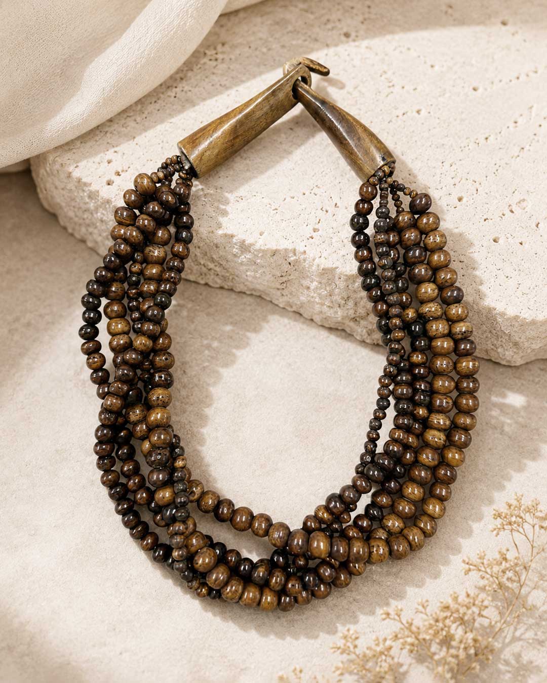 Brown five-strand beaded necklace laid on a textured stone surface with cream linen fabric and dried botanicals