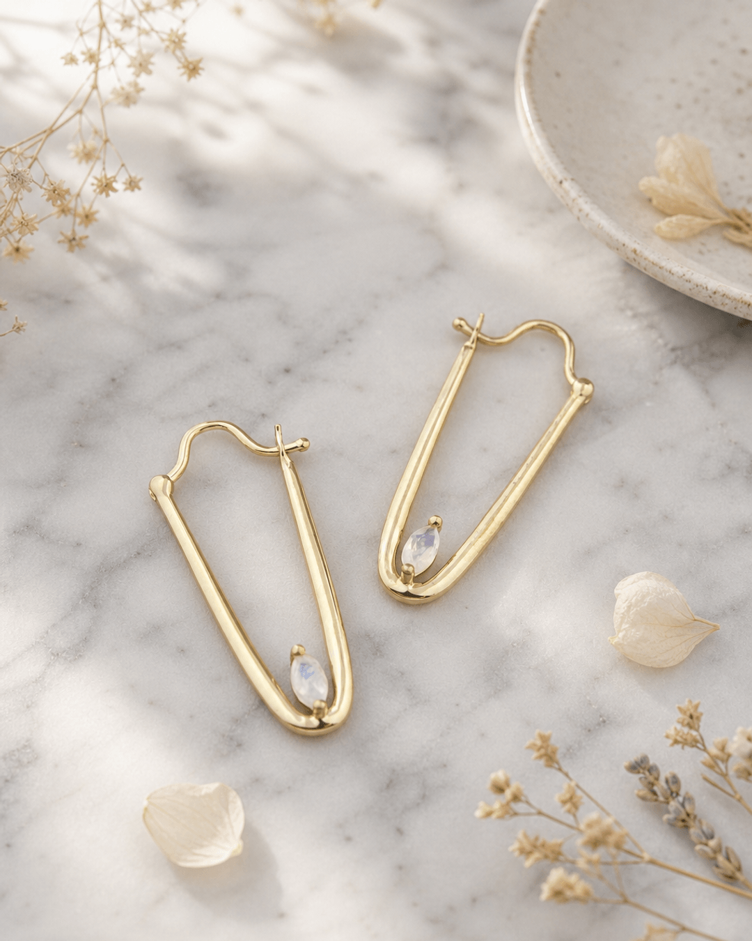 Gold plated moonstone drop earrings displayed on white marble surface, featuring iridescent oval gemstones and delicate gold settings
