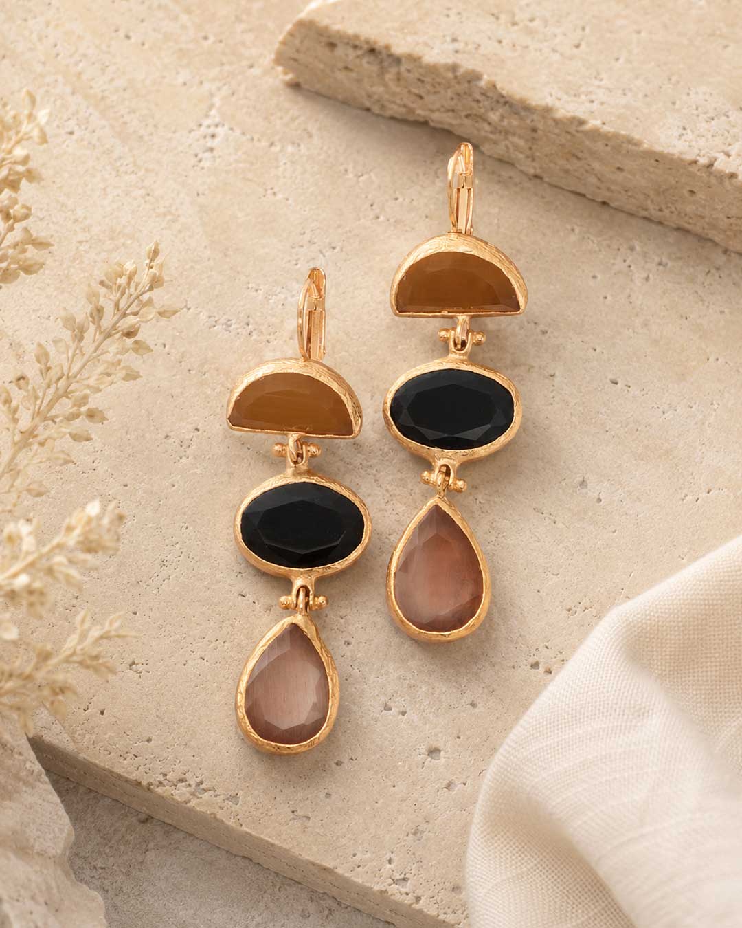 Gold-plated triple stone cat eye drop earrings on stone surface, flat lay showing amber, black, and blush stone set