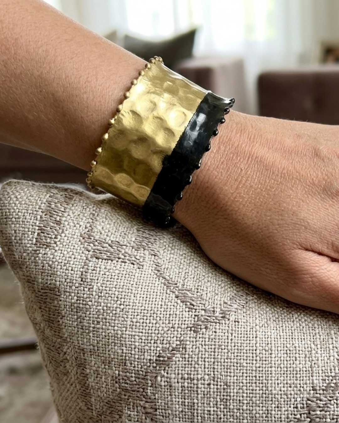 Hand-hammered brass cuff bracelet with black band on wrist resting on linen cushion, close-up showing hammered texture