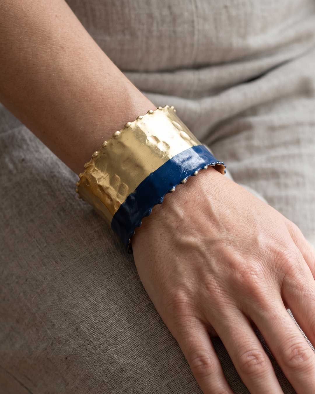 Hand-hammered brass cuff bracelet with indigo band worn on a wrist against a linen fabric background, angled side view
