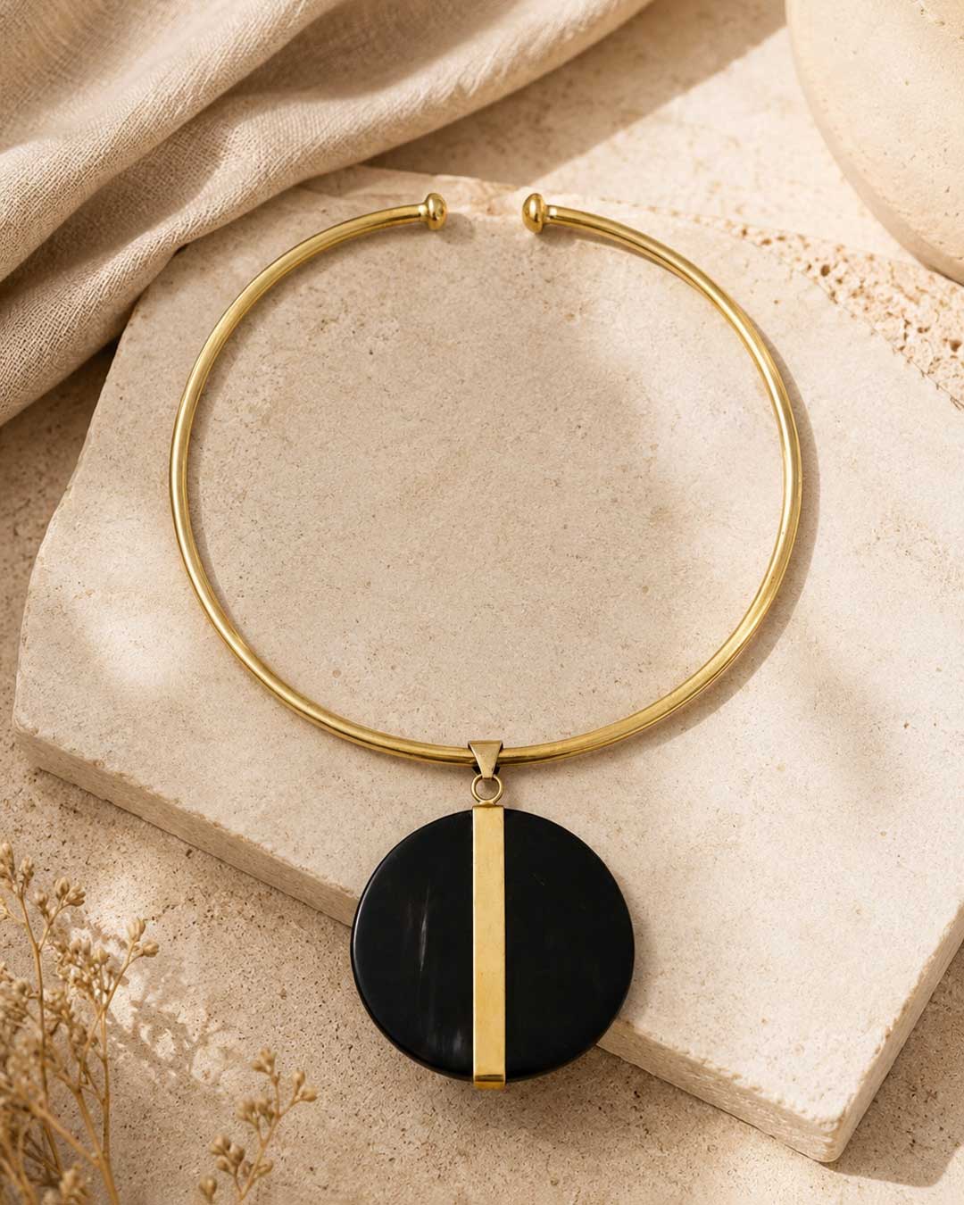 Horn and brass disc choker necklace styled on travertine stone surface with linen fabric and dried botanicals in background