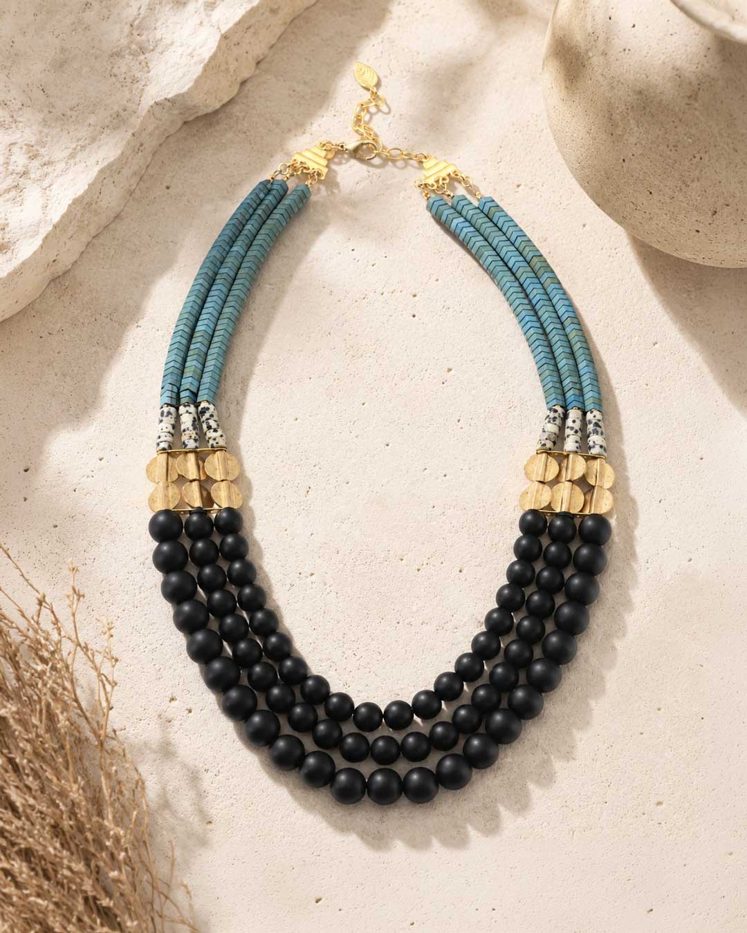 Jet glass and hematite triple strand necklace laid flat on stone surface with dried grass props, full layout and gold clasp visible