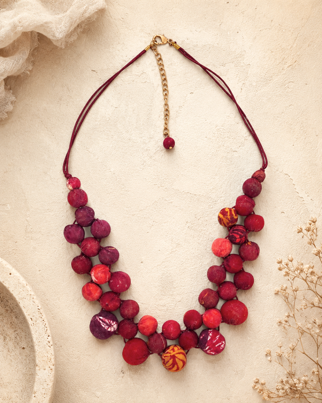 Kantha recycled textile layered bead necklace in red styled on a textured cream surface with dried botanicals alongside