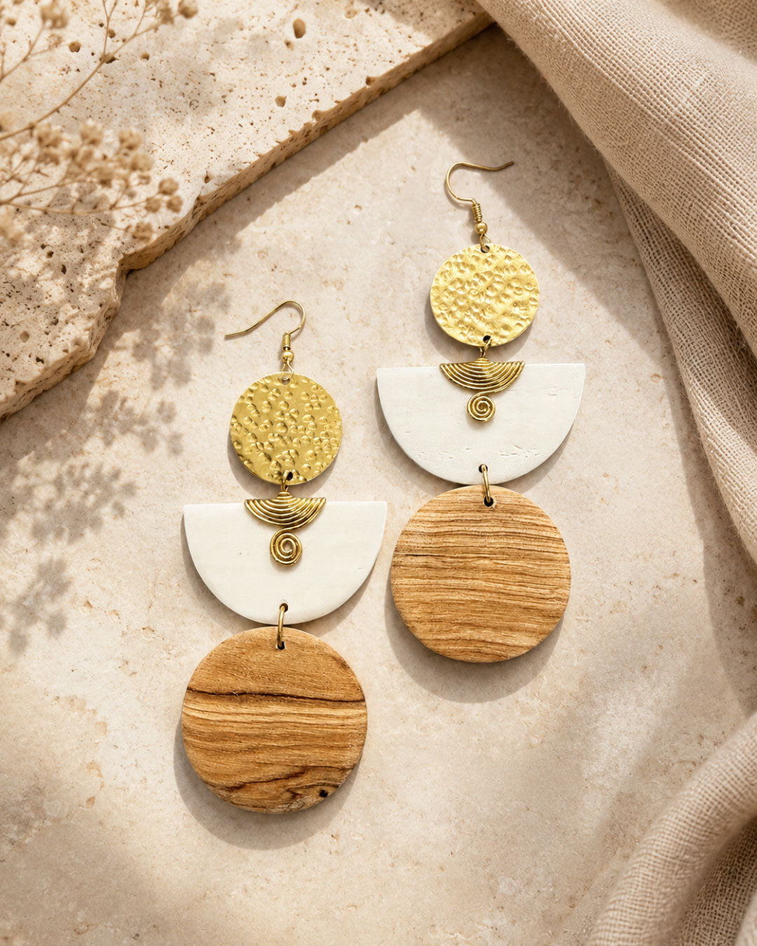 Kazuri brass and wood statement earrings laid flat on a textured stone surface alongside dried flowers and neutral linen fabric