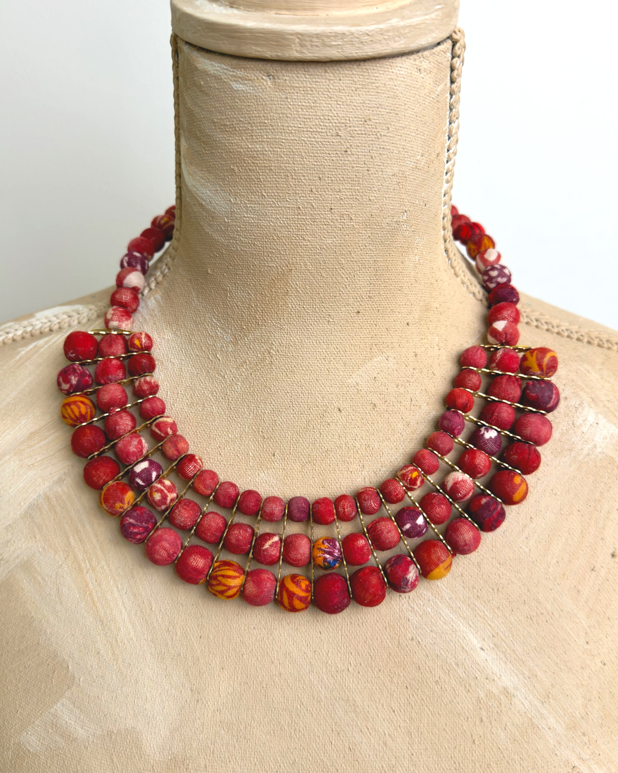 Red textile-wrapped bead layered bib necklace draped on a wooden dress form showing natural drape and mixed red bead rows