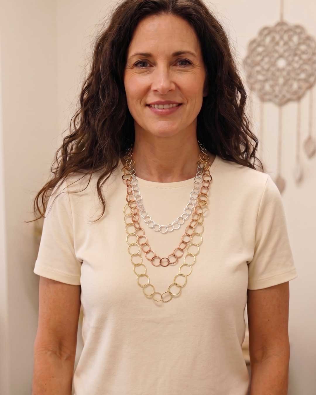 Three-strand mixed metal chain necklace worn by a woman in a cream t-shirt, showing layered gold, copper, and silver strands