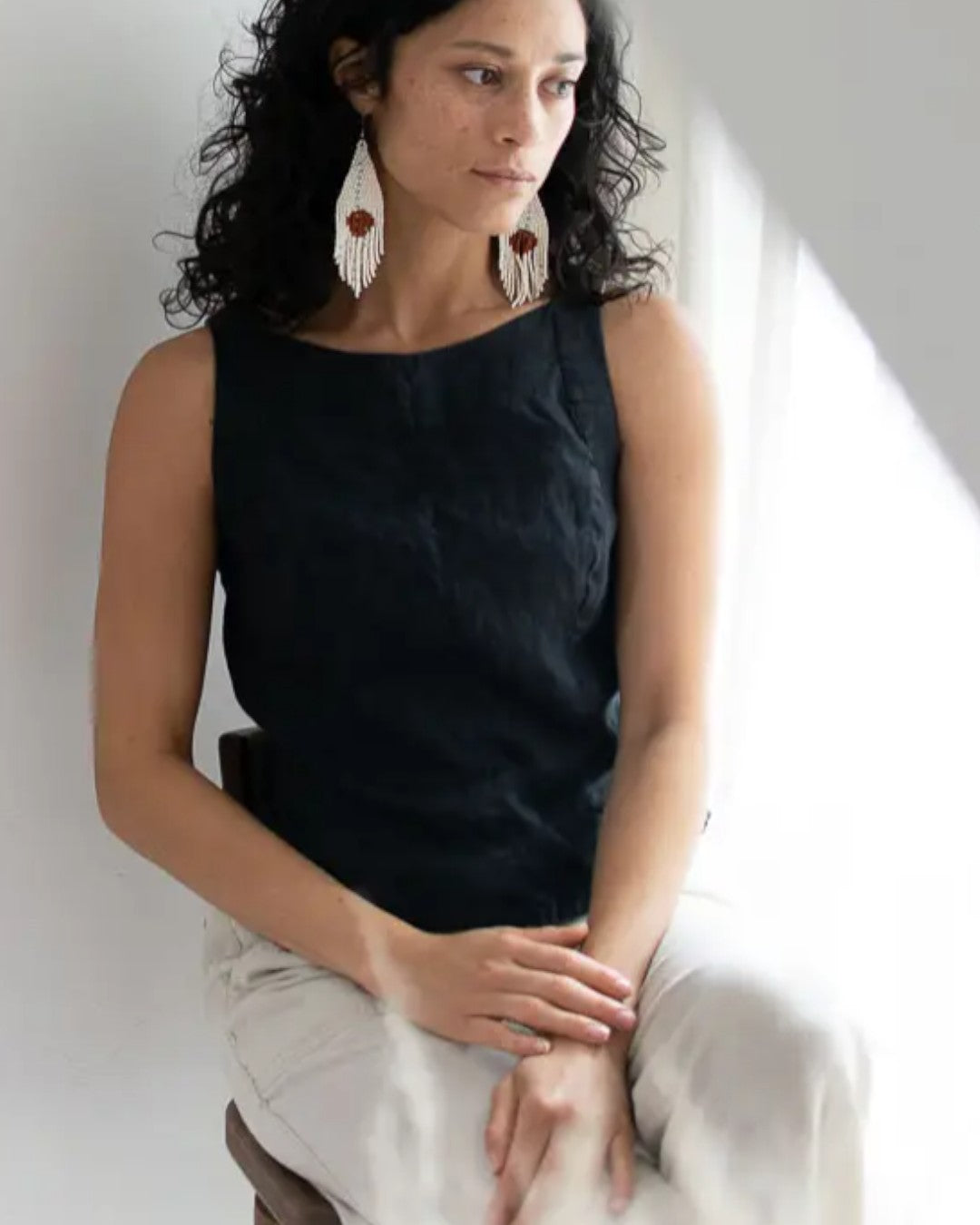 Woman wearing a black sleeveless top and white pants sitting against a neutral background
