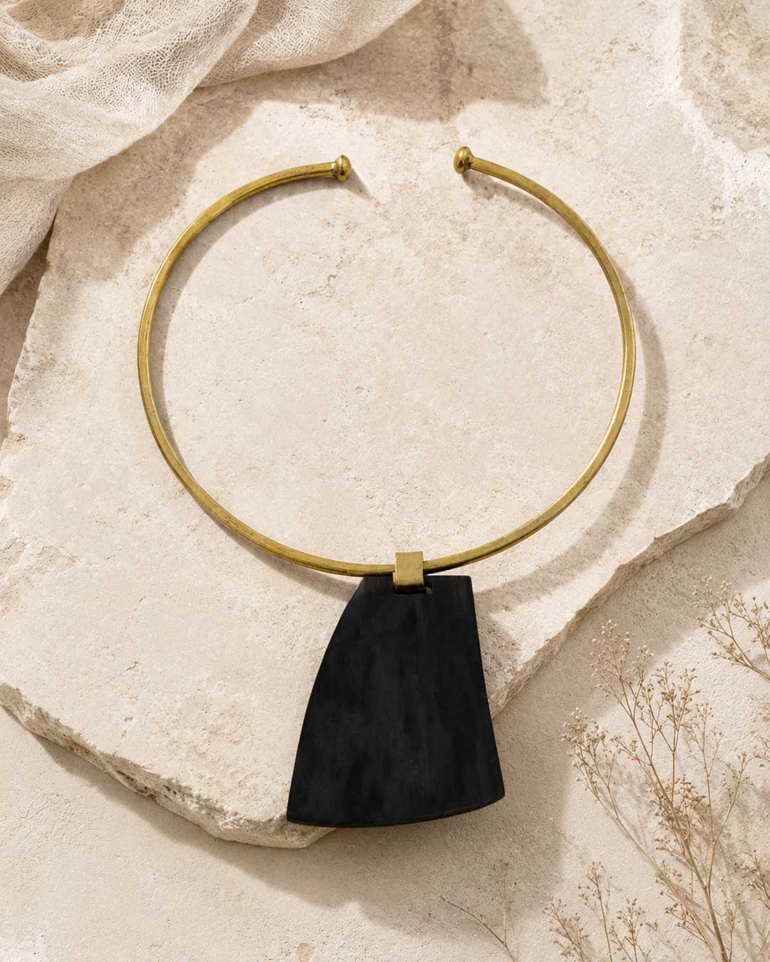 Ankole horn pendant brass choker necklace laid on textured stone surface with dried botanicals, showing full collar and pendant