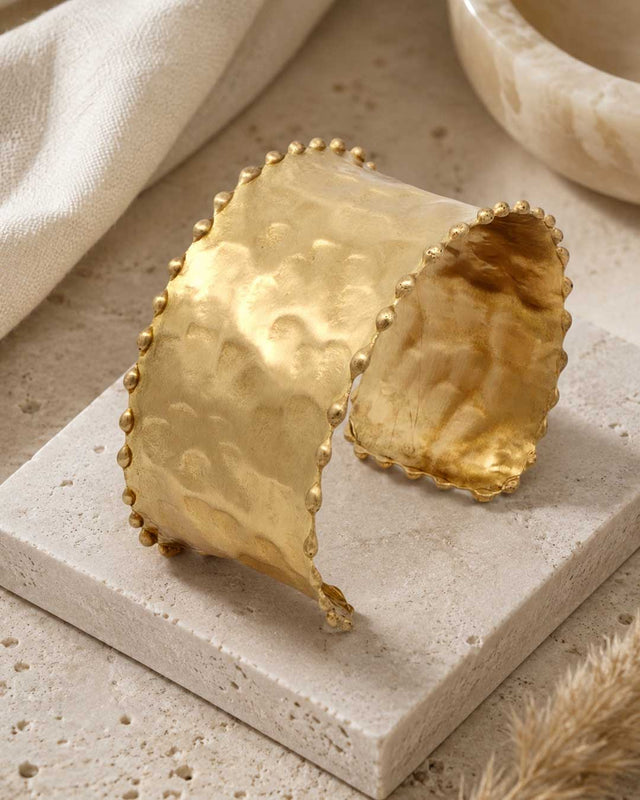 Beaded edge hand-hammered brass cuff bracelet standing upright on a stone tile, styled with linen cloth and dried grass