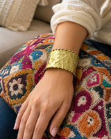 Beaded edge hand-hammered brass cuff bracelet worn on a wrist resting on a colorful woven pillow, worn view