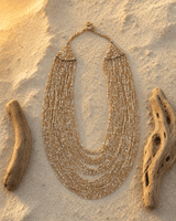 Beige and gold-tone multi-strand glass beaded necklace displayed on sandy pebble surface outdoors