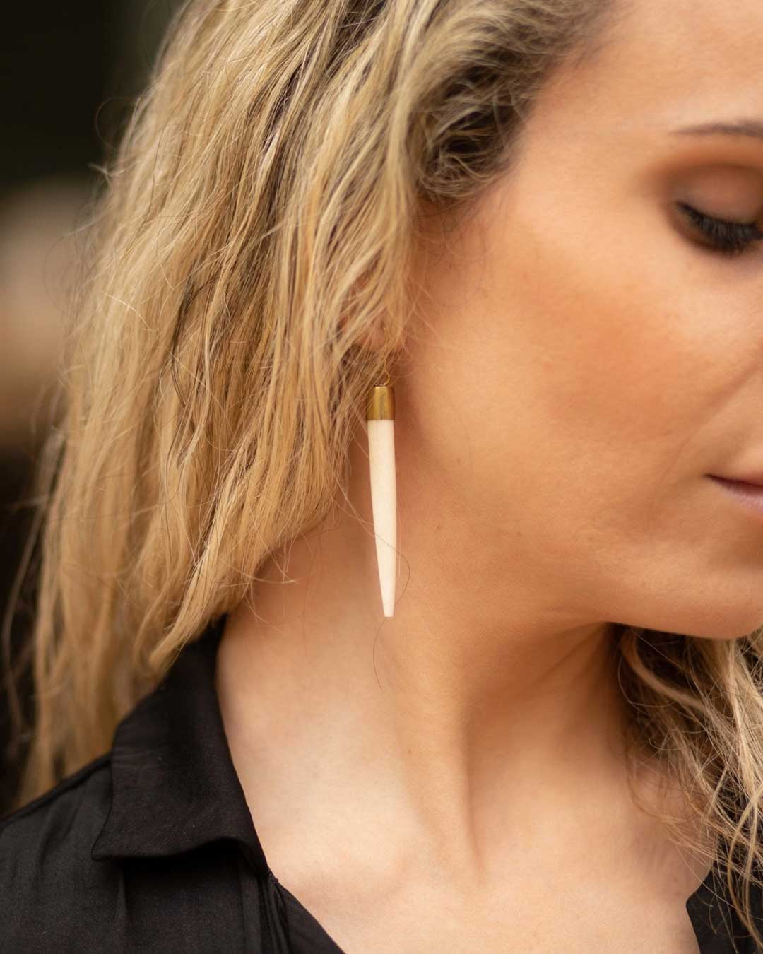 Bone and brass pencil spike dangle earring worn on a woman's ear, close-up side view against dark background