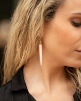 Bone and brass pencil spike dangle earring worn on a woman's ear, close-up side view against dark background