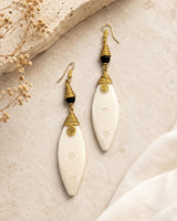 Kenyan hand-coiled brass and carved bone drop earrings styled on stone surface with dried flowers and white linen fabric
