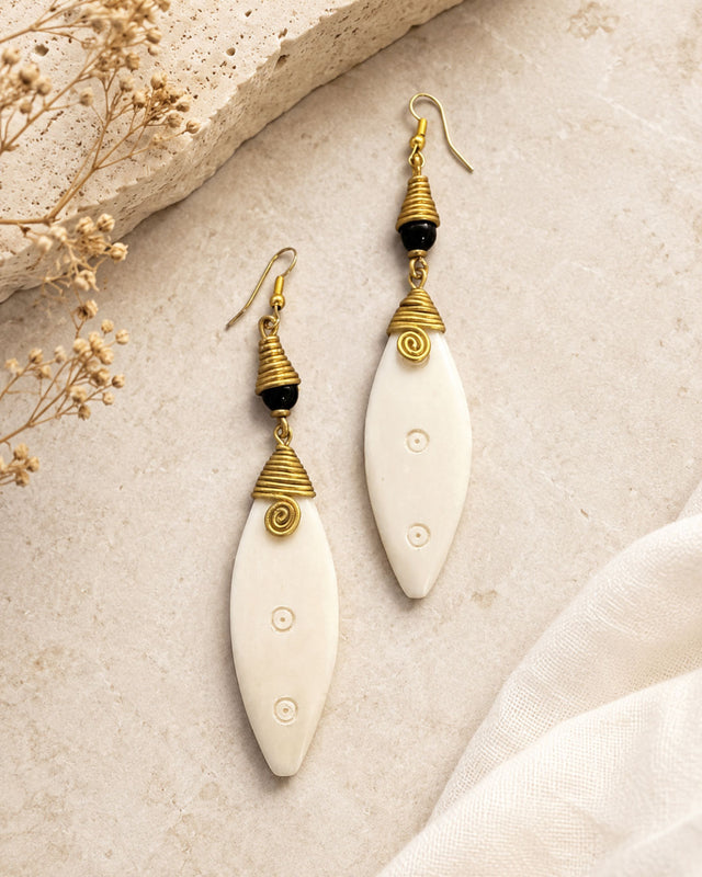 Kenyan hand-coiled brass and carved bone drop earrings styled on stone surface with dried flowers and white linen fabric