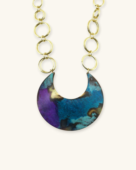 Hand-patinated brass crescent pendant necklace close-up showing blue, teal, and purple patina with gold chain links