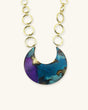 Hand-patinated brass crescent pendant necklace close-up showing blue, teal, and purple patina with gold chain links