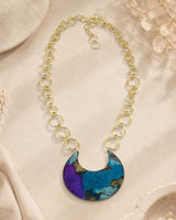 Hand-patinated brass crescent pendant necklace arranged flat on a textured beige surface with dried botanical props nearby