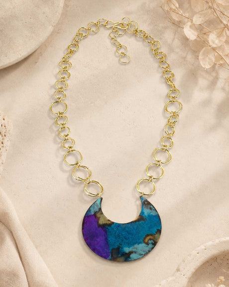 Hand-patinated brass crescent pendant necklace arranged flat on a textured beige surface with dried botanical props nearby