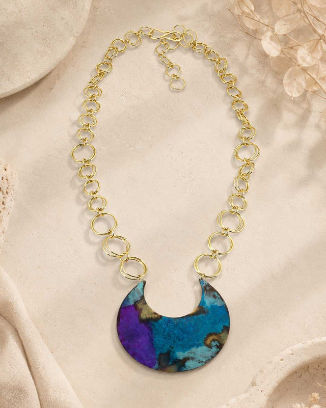 Hand-patinated brass crescent pendant necklace arranged flat on a textured beige surface with dried botanical props nearby