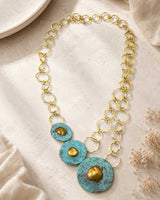 Hand-forged brass disc blue patina necklace laid flat on stone surface showing toggle clasp, double ring chain, and three graduated discs