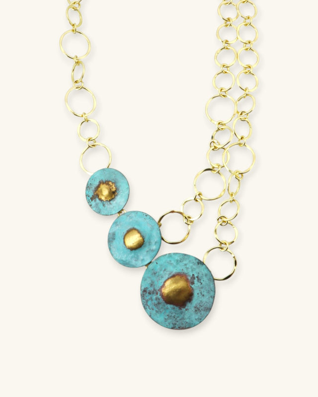 Hand-forged brass disc necklace showing three graduated blue patina discs with gold centers on open-link chain, angled close-up view