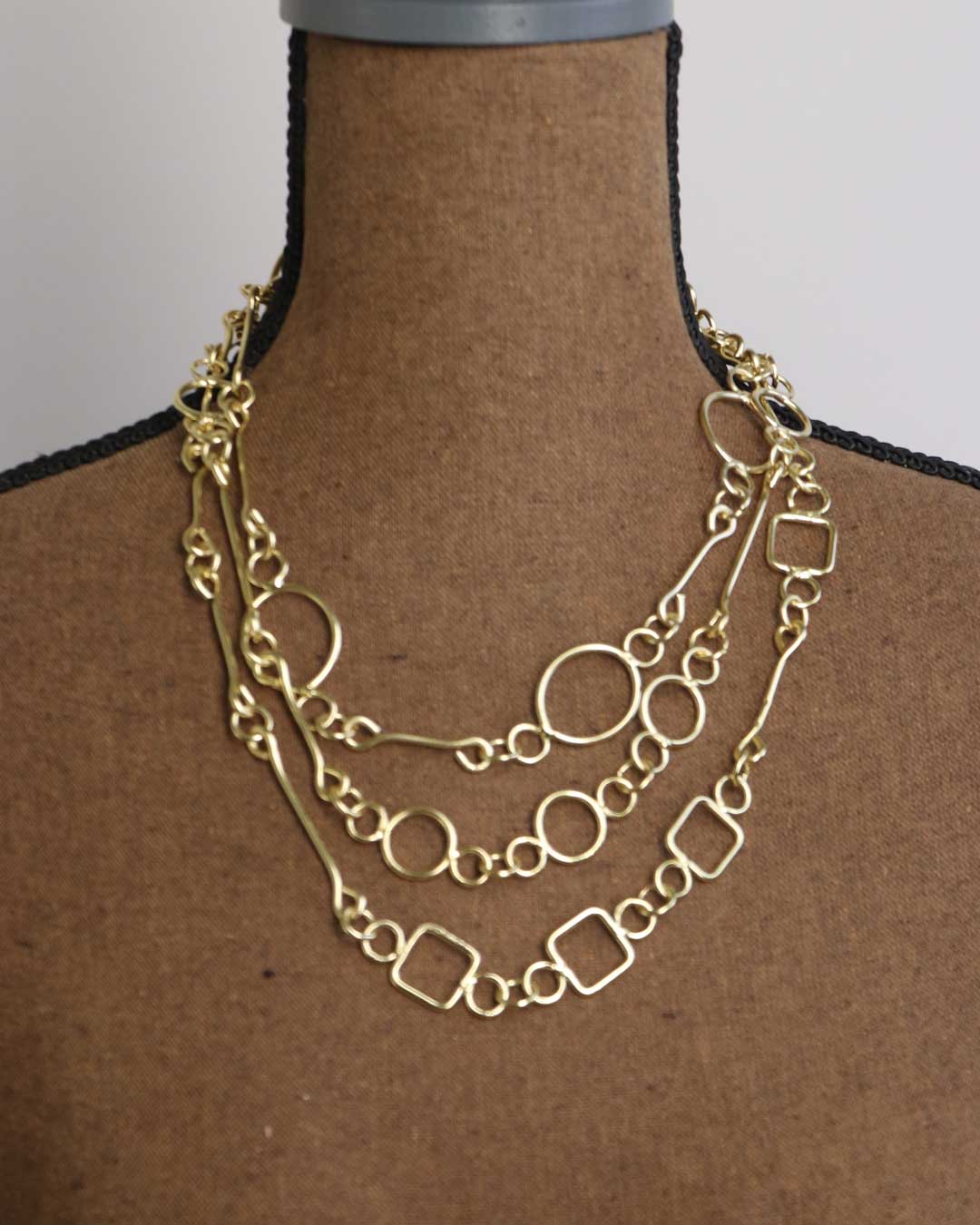 Brass link geometry long wrap necklace layered as three strands on dress form neck, showing circular and square link detail