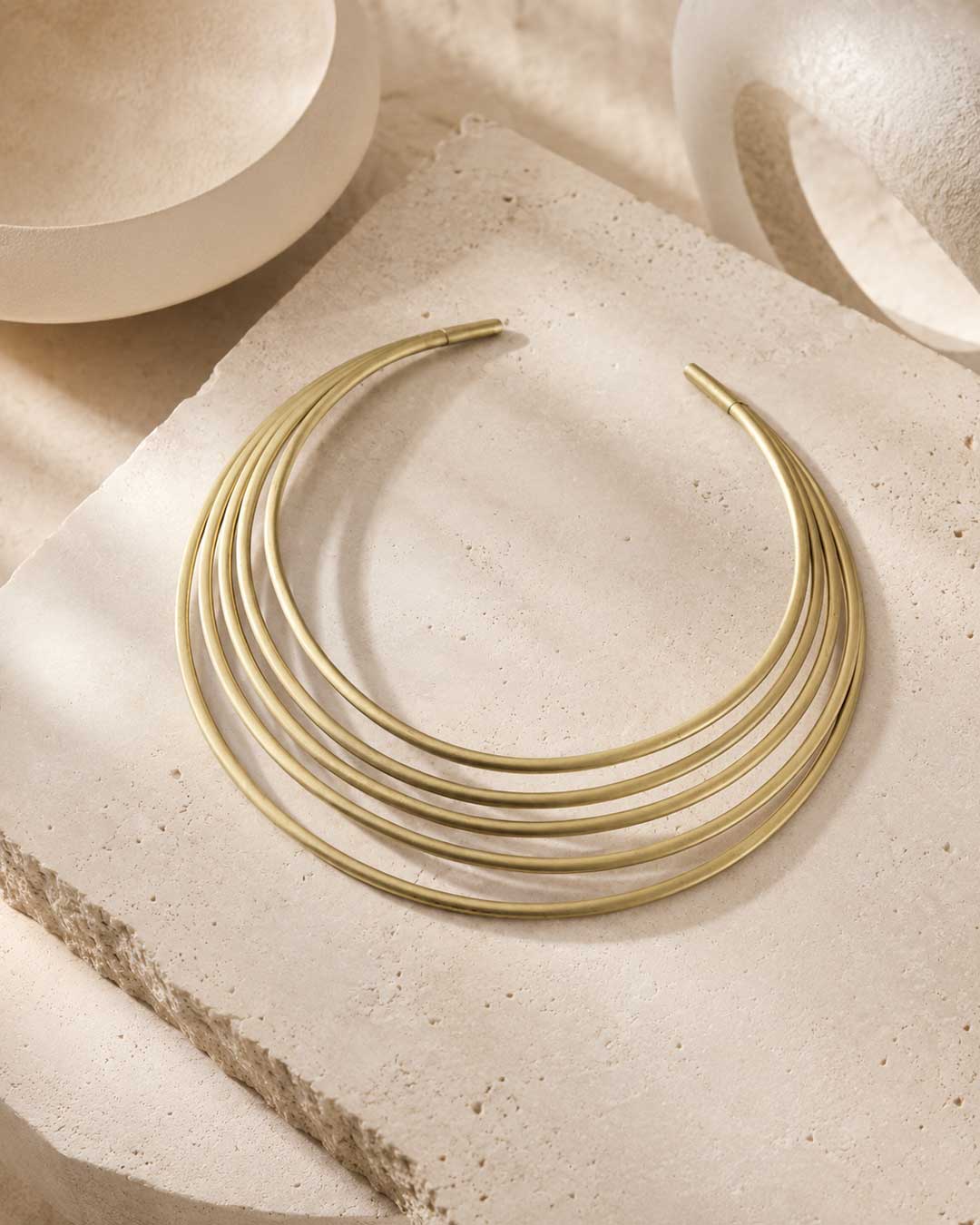 Brass multi-band collar necklace laid flat on a rough stone surface with ceramic bowls and draped linen fabric