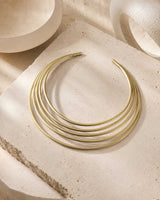 Brass multi-band collar necklace laid flat on a rough stone surface with ceramic bowls and draped linen fabric