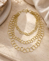 Brass three-strand circular link chain necklace flat lay on stone surface with toggle clasp visible, warm styled shot