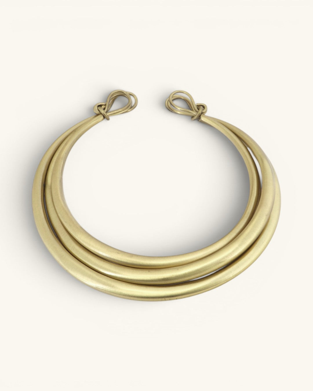 Hand-beaten brass triple-strand collar necklace on cream background, front view showing wire loop closures at each end