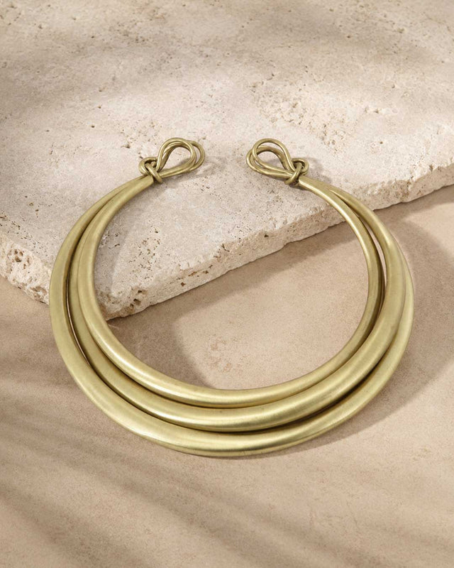 Hand-beaten brass triple-strand collar necklace laid on sandy travertine stone, overhead view showing clasp ends