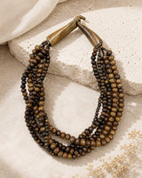 Brown five-strand beaded necklace laid on a textured stone surface with cream linen fabric and dried botanicals
