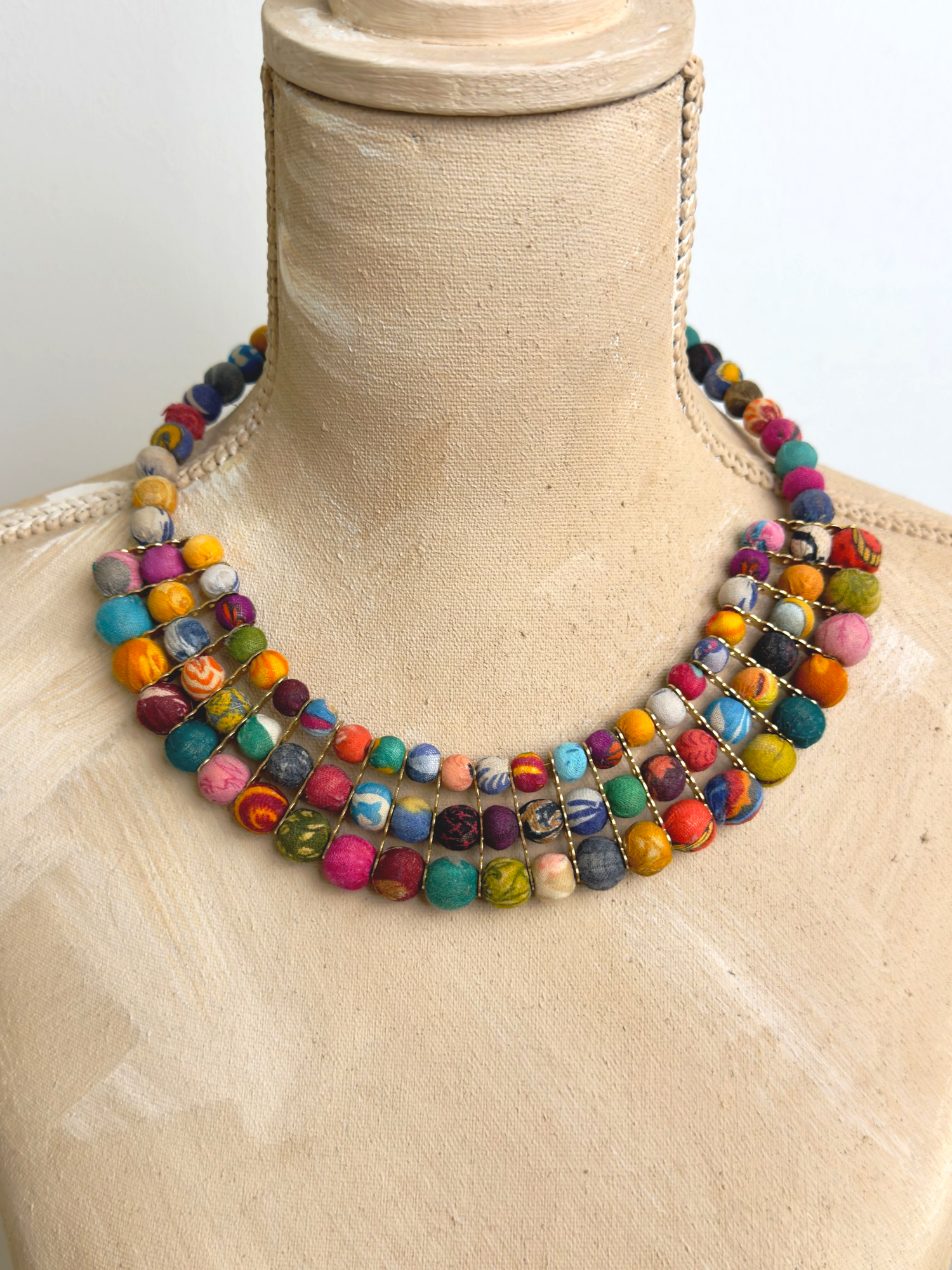 Colorful textile-wrapped bead layered necklace displayed on a tan fabric dress form, showing three-row bib drape