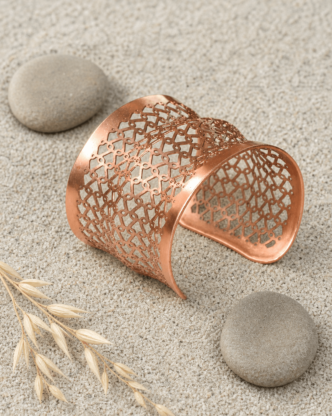 Copper wide cuff bracelet with geometric lattice pattern resting on sandy pebbles outdoors