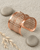 Copper wide cuff bracelet with geometric lattice pattern resting on sandy pebbles outdoors
