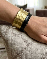 Hand-hammered brass cuff bracelet with black band on wrist resting on linen cushion, close-up showing hammered texture