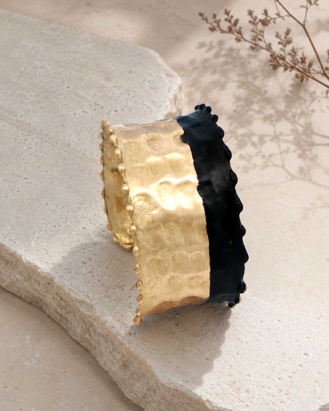 Hand-hammered brass cuff bracelet with black band resting on stone surface, angled view showing gold and black contrast