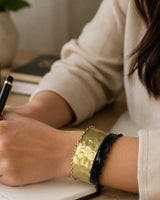 Hand-hammered brass cuff bracelet with black band worn on wrist, lifestyle shot with notebook and pen in background