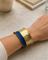 Hand-hammered brass cuff bracelet with indigo band worn on a wrist, top-down view showing hammered texture and beaded edge detail