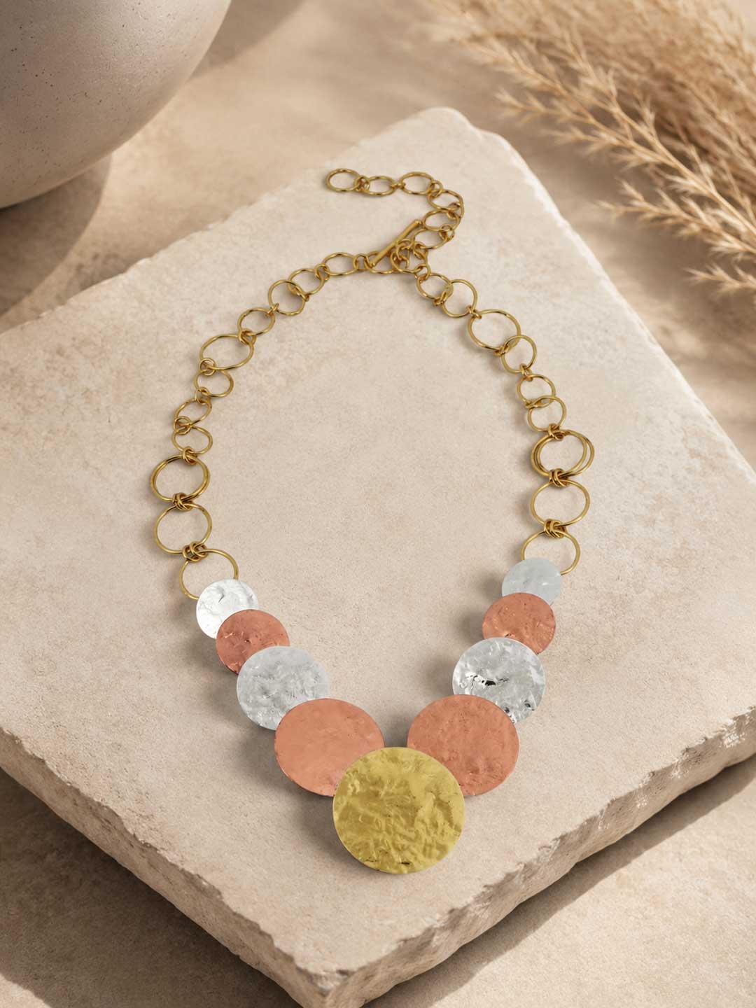 Hammered tri-tone disc necklace laid flat on a sand-colored stone surface with pampas grass, showing full length and clasp detail