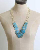 Hand-forged brass statement necklace with blue patina discs, primary front view displaying the complete necklace design