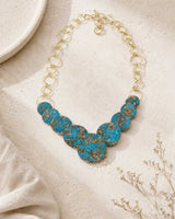 Hand-forged brass statement necklace with blue patina discs, front angle showing full length chain and pendant arrangement