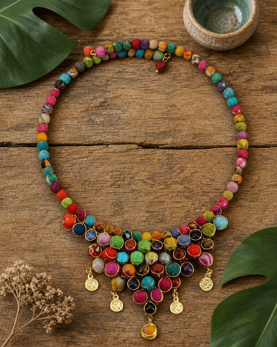 Handmade multicolor textile and gemstone choker necklace displayed on a flatlay with green botanical leaves