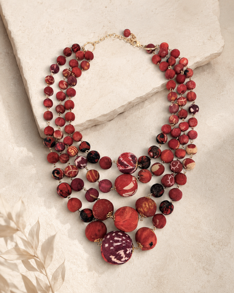 Four-strand kantha sari bead necklace in red tones laid flat on a stone surface with a dried botanical prop
